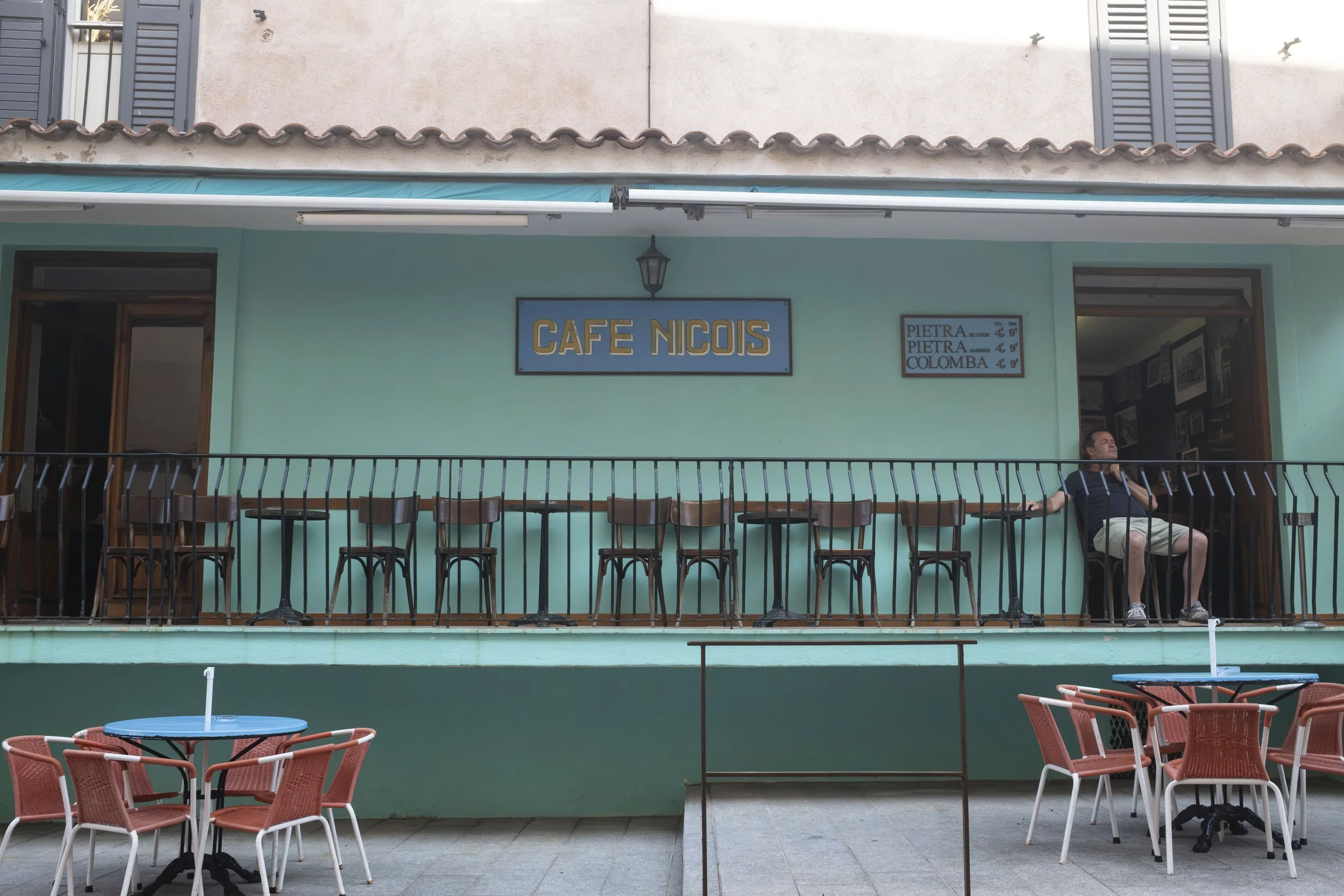 Façade verte d'un café niçois, un homme est assis sur une chaise au balcon, en premier plan, en terrasse, les chaises en plastiques rouges et les tables bleues.