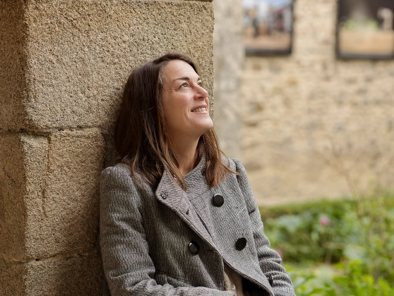 Femme souriante posant contre un mur en pierre, regardant vers le haut, en extérieur près d'une bâtisse en pierre et d'un jardin.