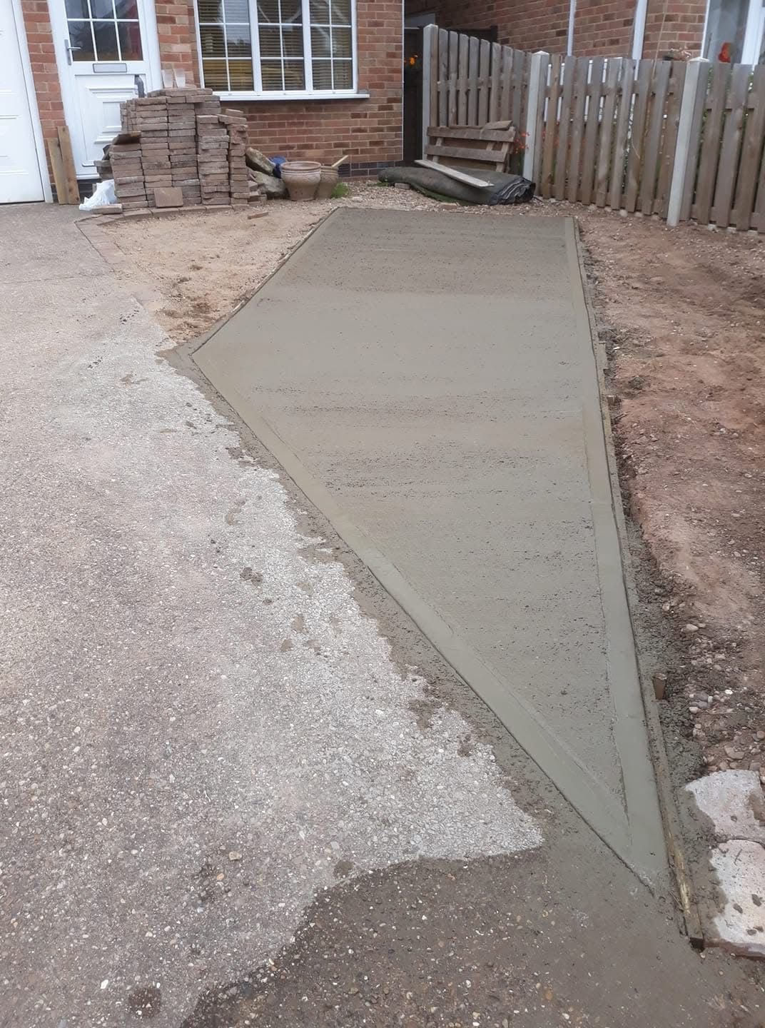 Freshly poured concrete patio next to a brick house with a stack of tiles nearby.