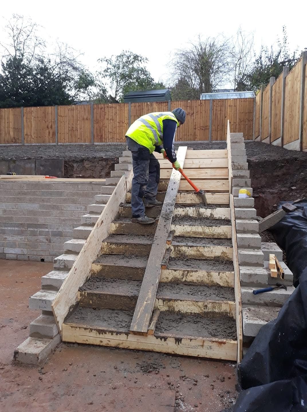 Construction worker in high-vis vest building concrete stairs with wooden formwork outdoors.