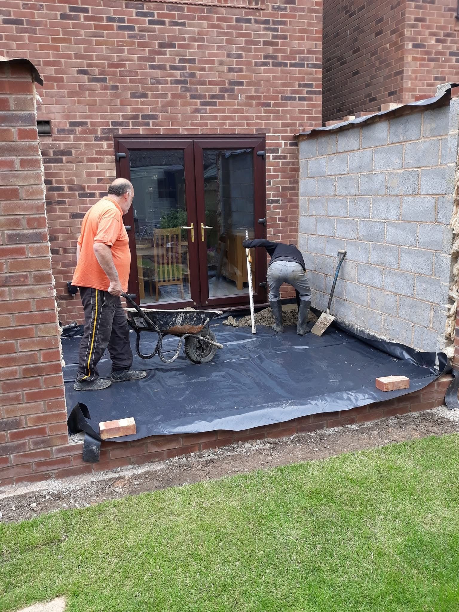 Two men working on building a small extension with brick walls, using a wheelbarrow and tools, near a house with a glass door.