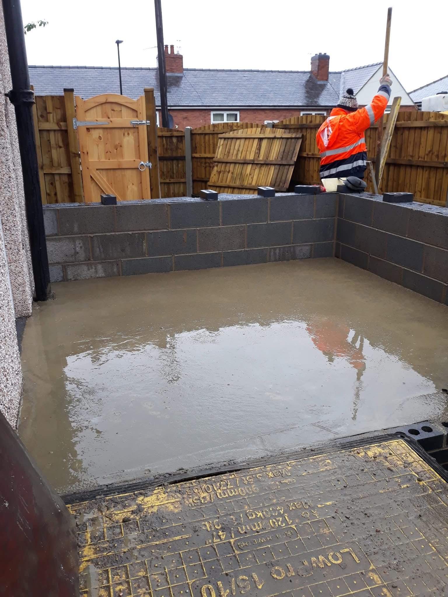 Construction site with fresh concrete foundation, brick walls, and a worker in an orange jacket adjusting a post. Wooden fence and gate in the background.