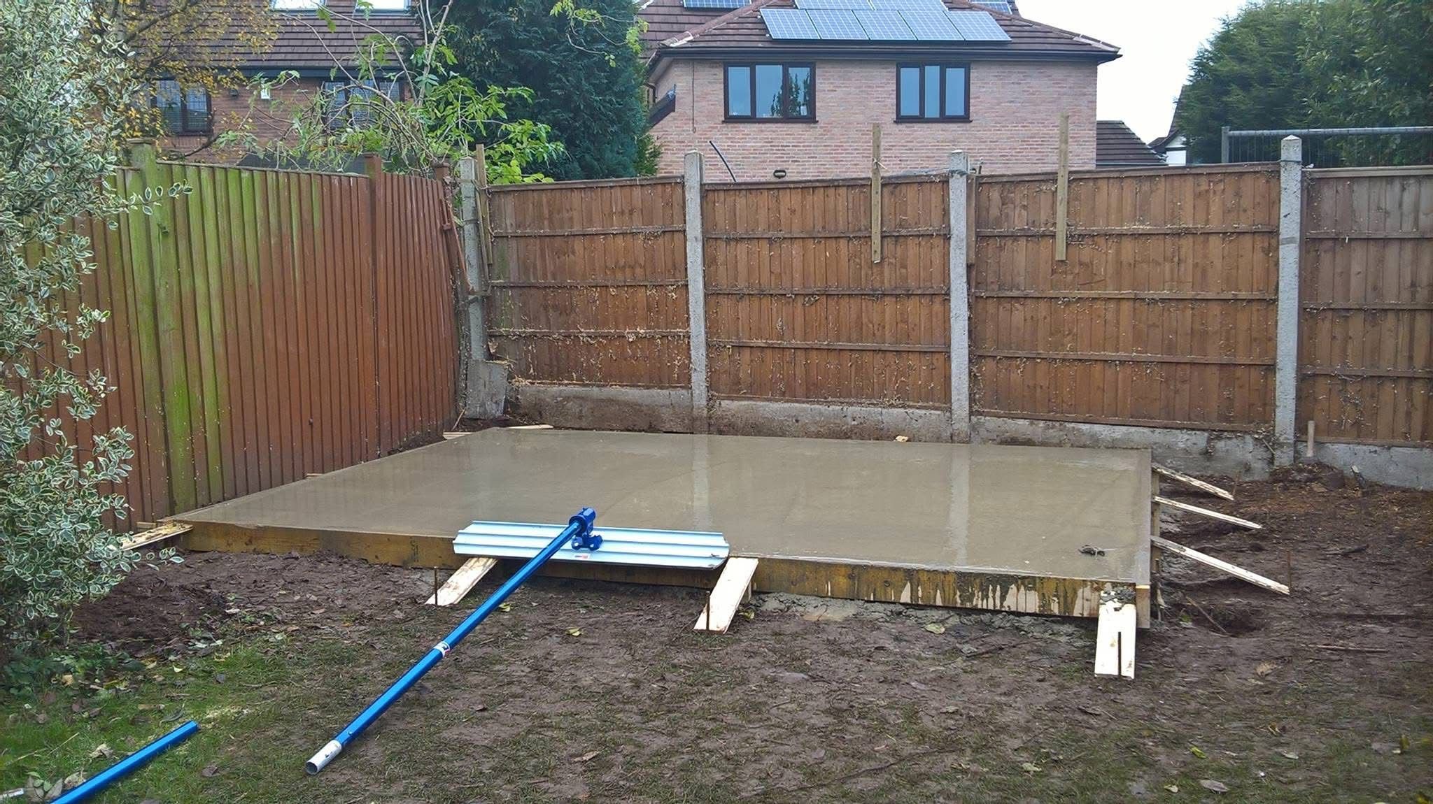 Freshly poured concrete slab in a backyard with wooden formwork and a float tool, surrounded by fences and grass.