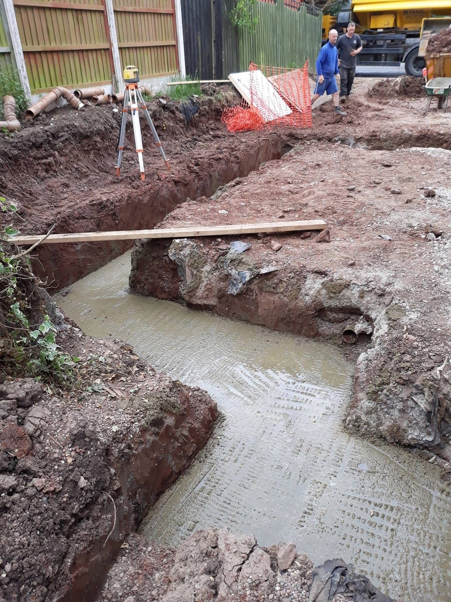 Construction site with freshly poured concrete foundation trenches and a laser level on a tripod, surrounded by soil, pipes, and construction materials. Two workers are visible in the background.