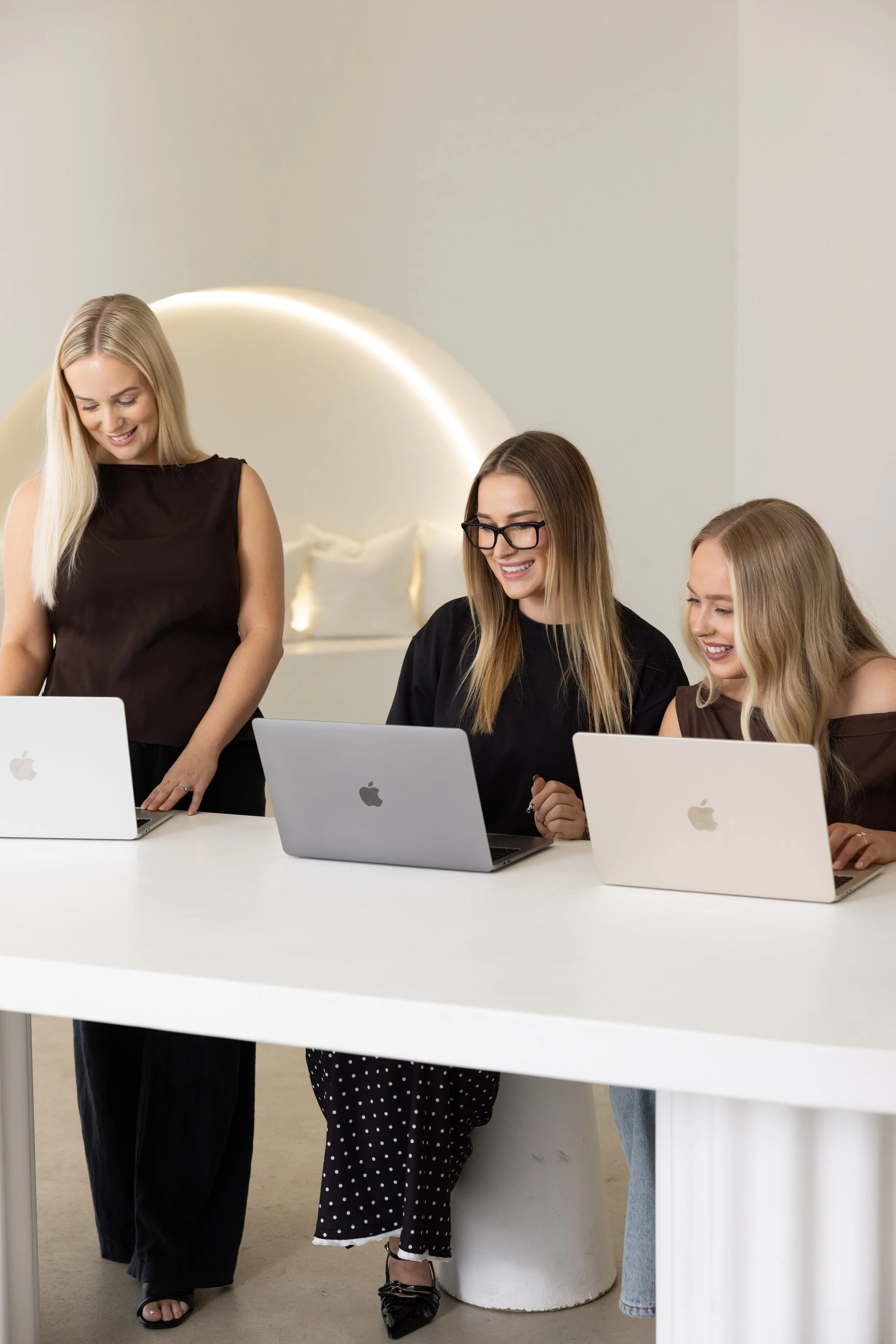 Four women in an office or conference room, three sitting at a white desk with laptops and one standing, smiling and engaging with the seated women.
