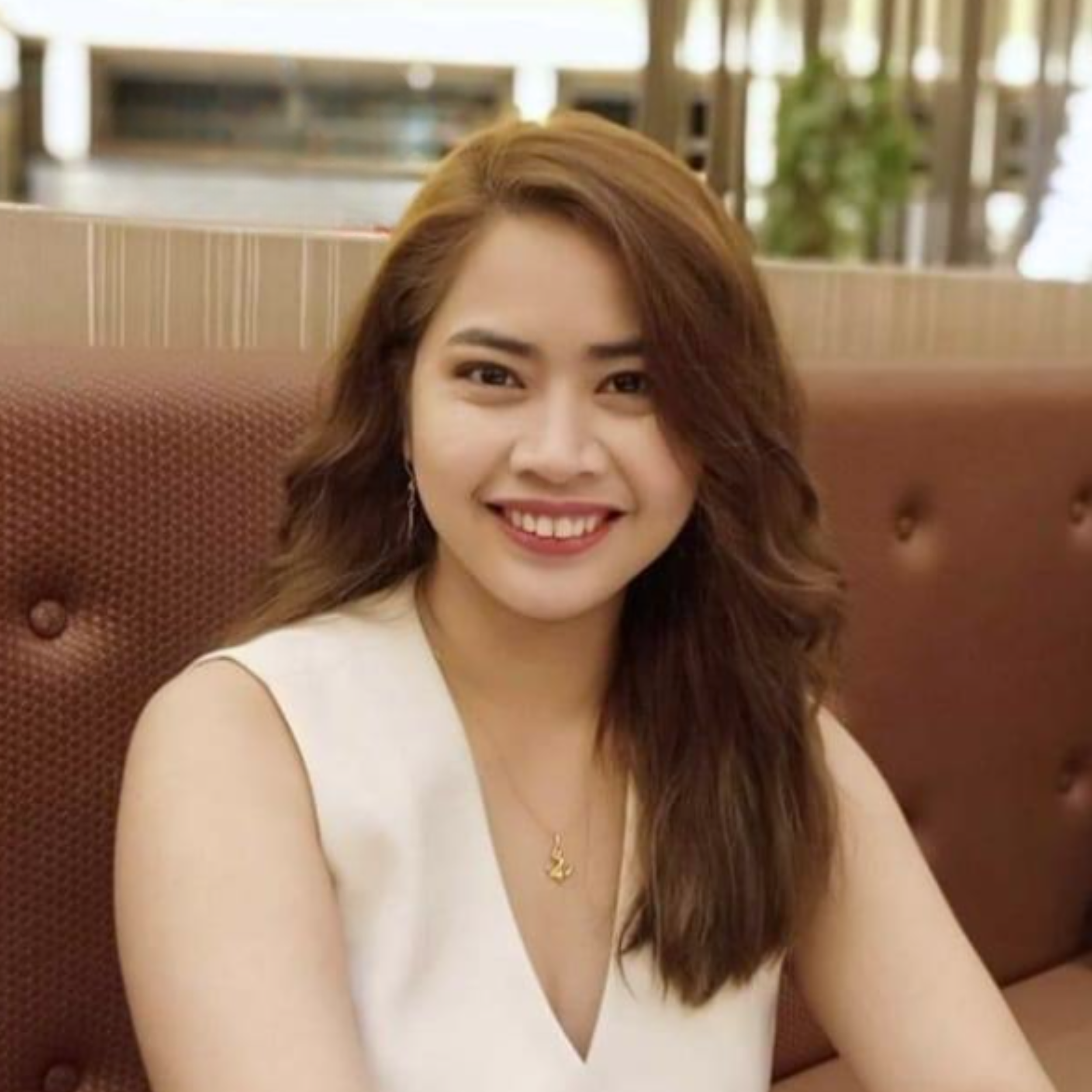 Close-up of a woman with long brown hair, smiling, wearing a white sleeveless top and a gold necklace, sitting in a restaurant booth.