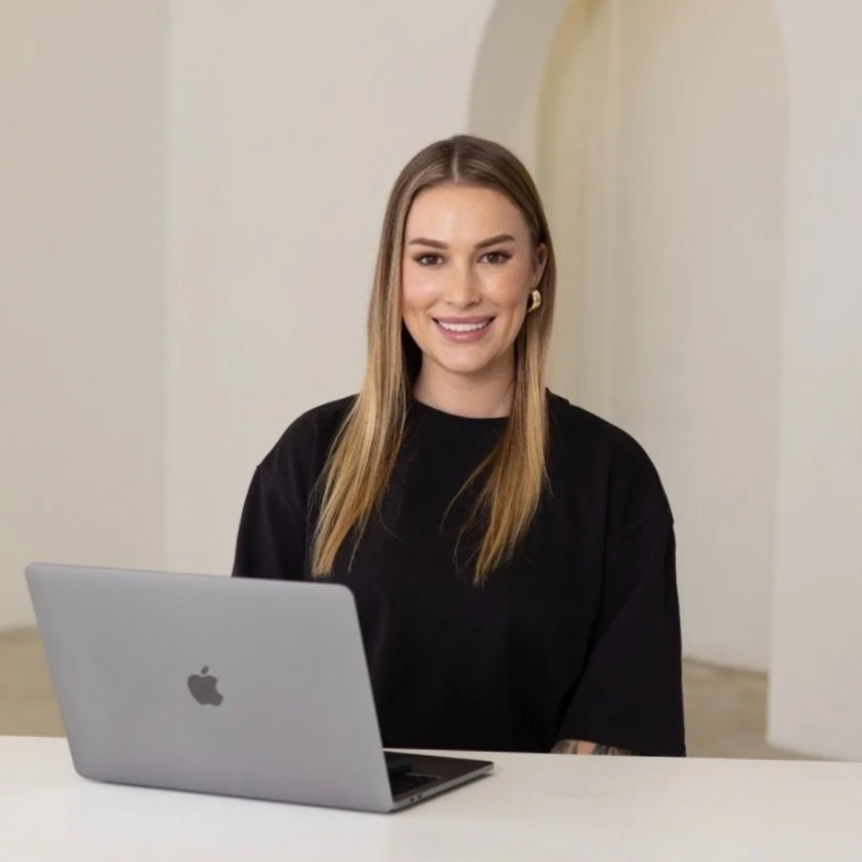 A woman with long blonde hair smiling at the camera, sitting at a white table with a silver MacBook laptop in front of her, in a well-lit room with white walls.