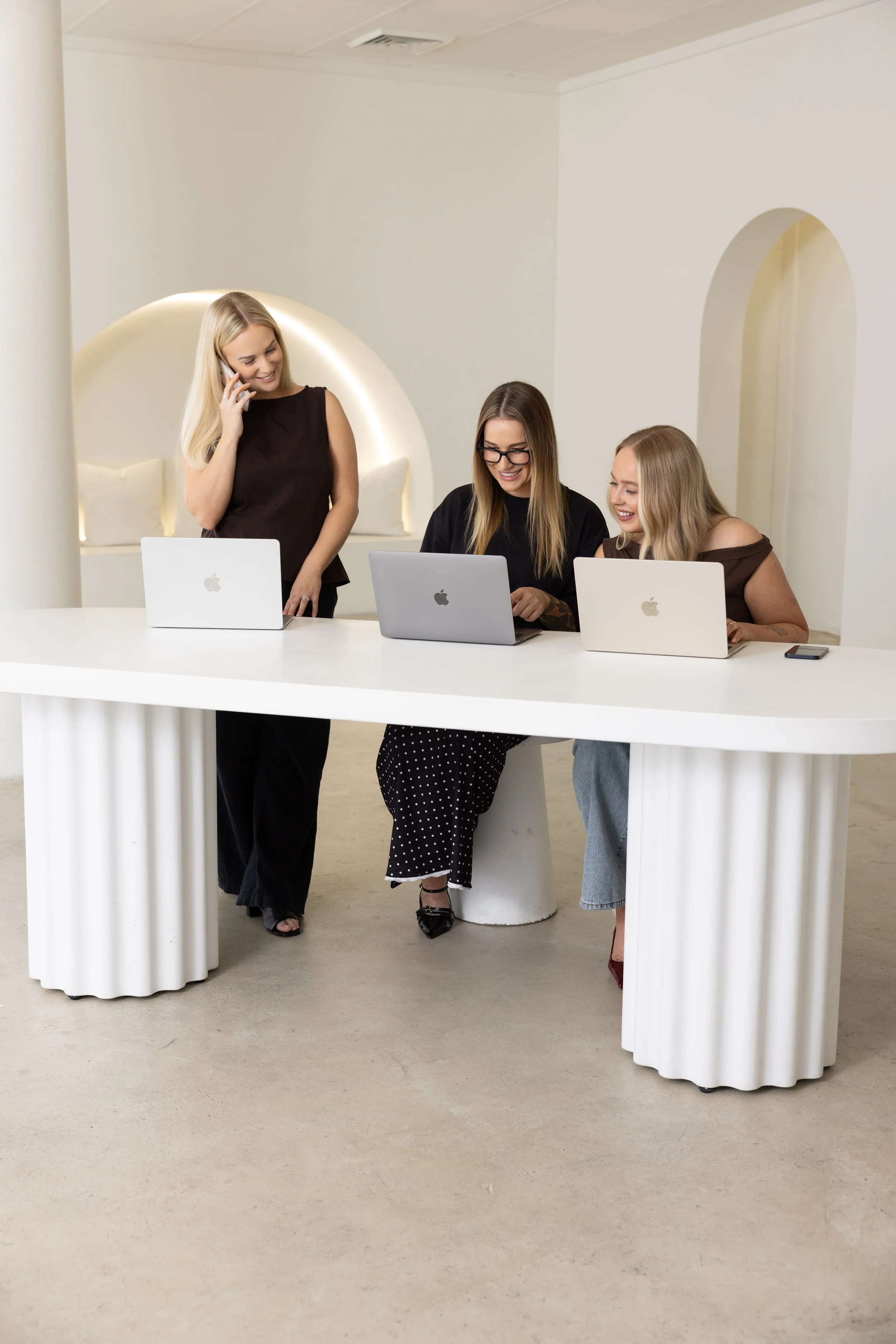 Three women with laptops at a white table, two seated and one standing, smiling and engaged in a conversation in a modern, minimally decorated room.