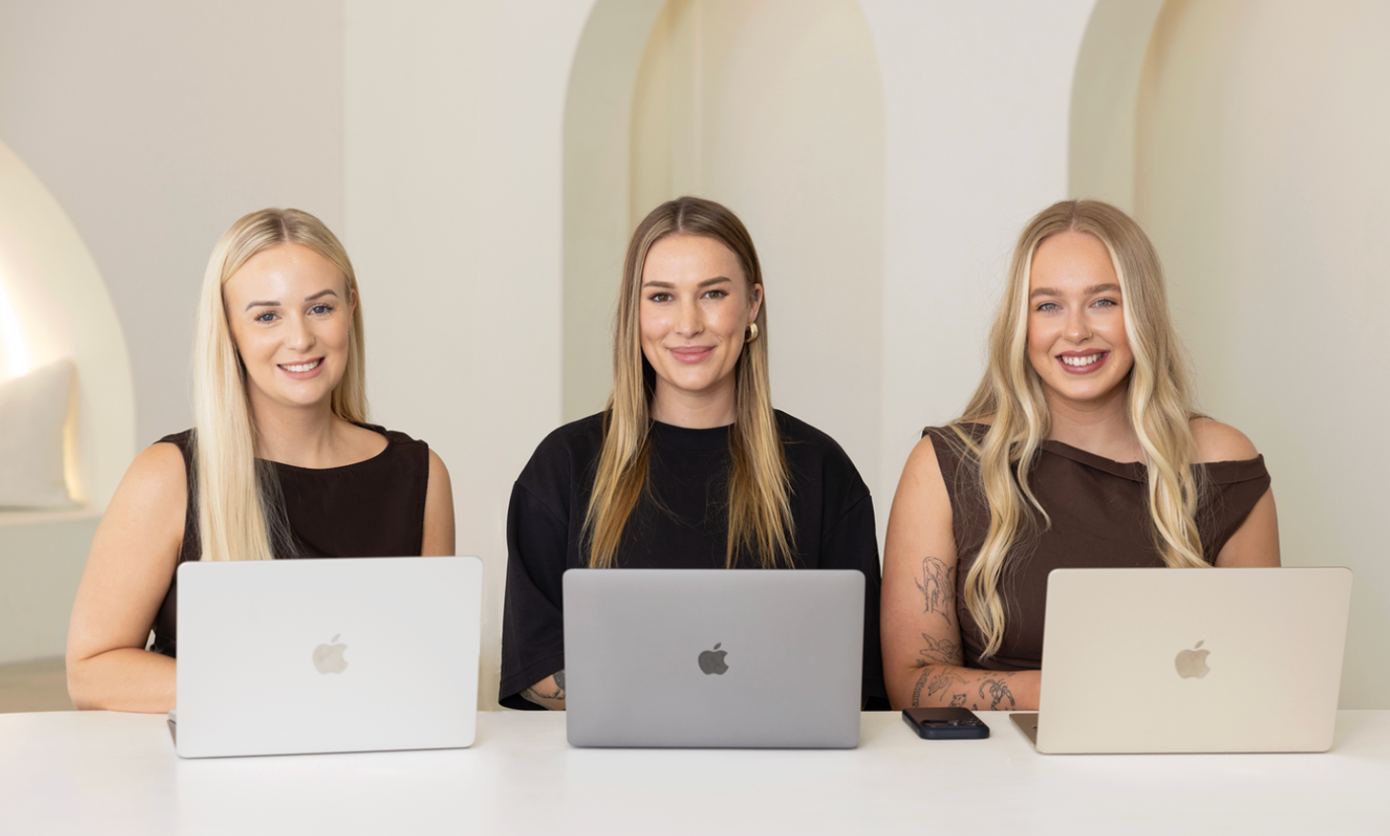 Three women sitting at a table with laptops, smiling at the camera, in a bright room with white walls.