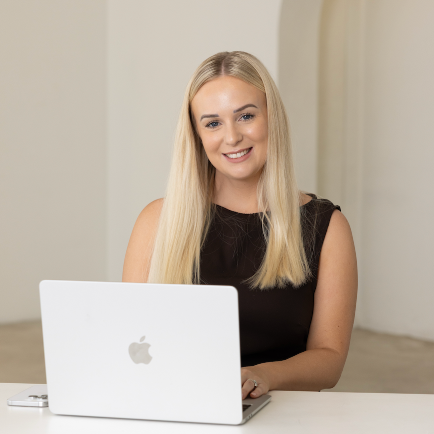 A woman with long blonde hair smiling while sitting at a table with an open Apple MacBook laptop.