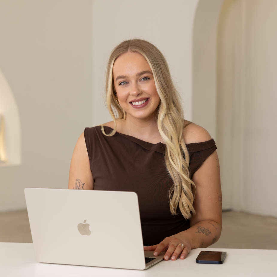 Smiling woman with long blonde hair sitting at a table with a white laptop and a smartphone.