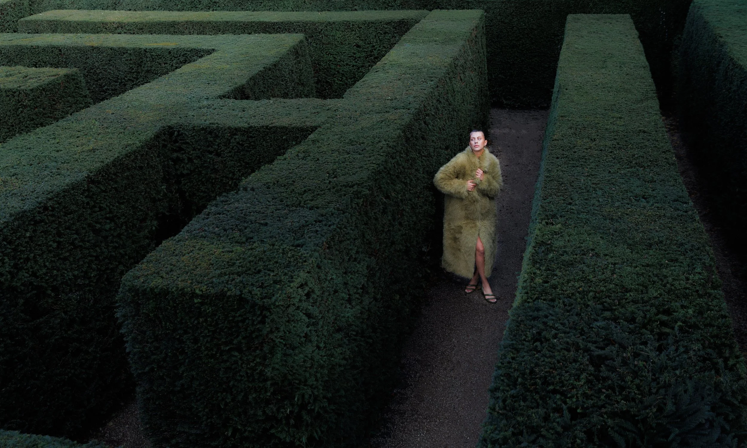A woman standing in a hedge maze, wearing a long fur coat with a slit, and sandals, looking upwards.