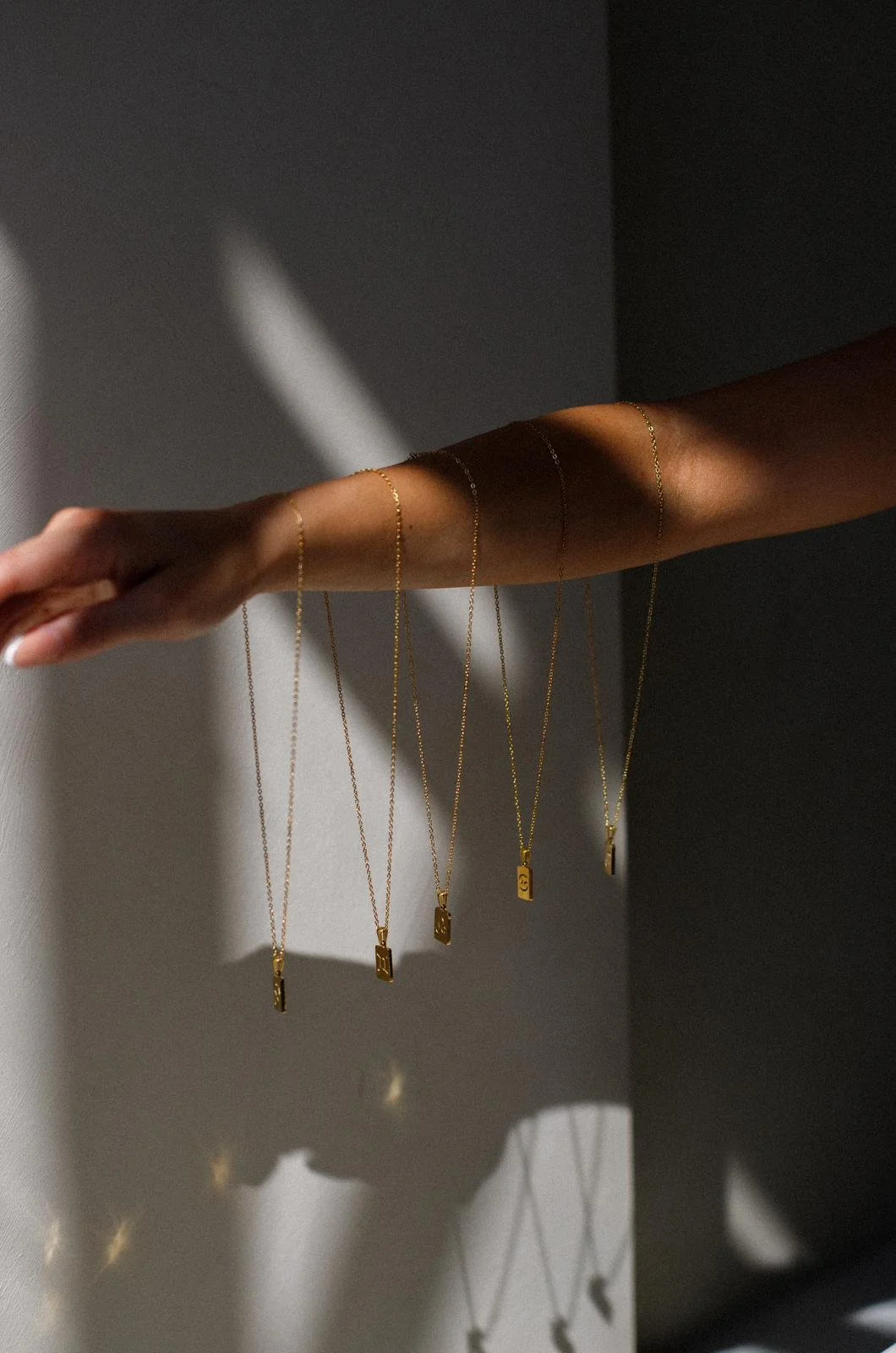 A person’s arm extended horizontally with several gold necklaces hanging from their arm, casting shadows on a white wall.