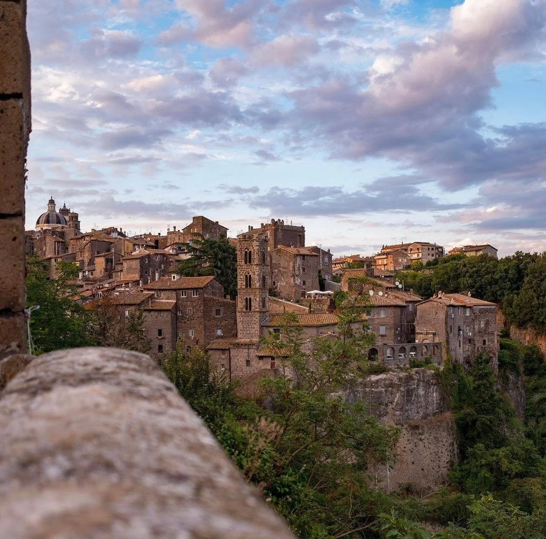 Panorama di un borgo medievale italiano al tramonto, con edifici in pietra su una collina e cielo nuvoloso sullo sfondo.