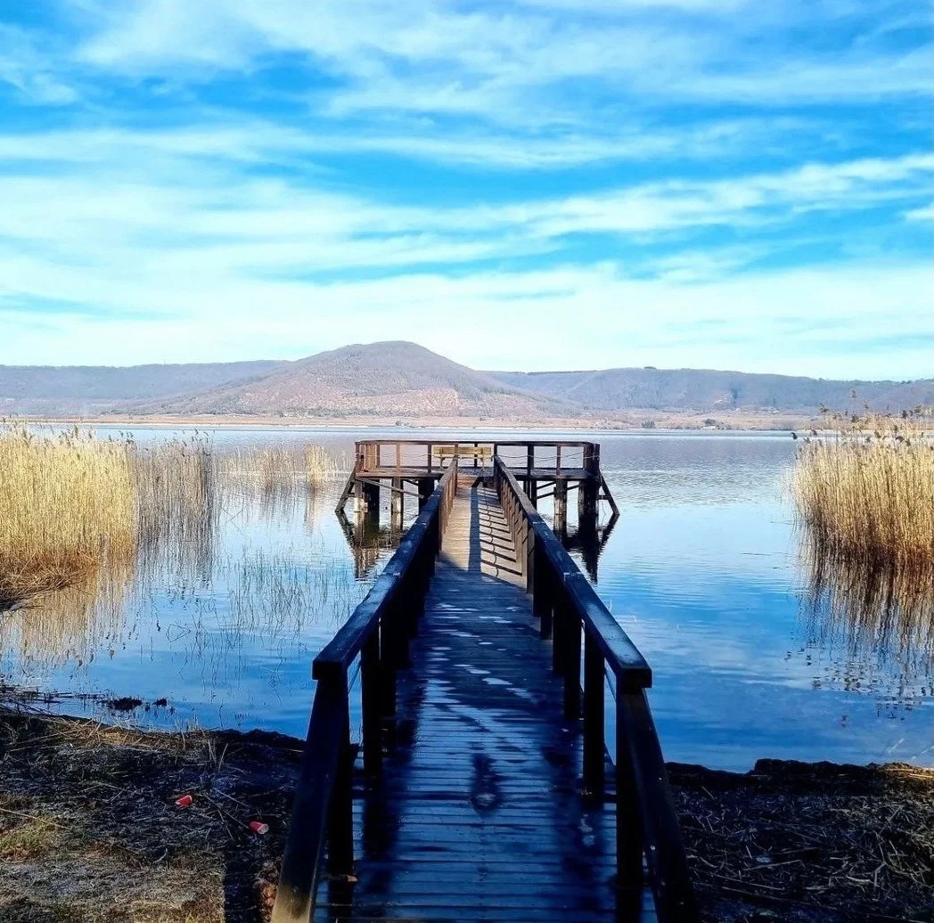 Lago con pontile di legno, canne palustri ai lati e una montagna sullo sfondo sotto un cielo sereno.
