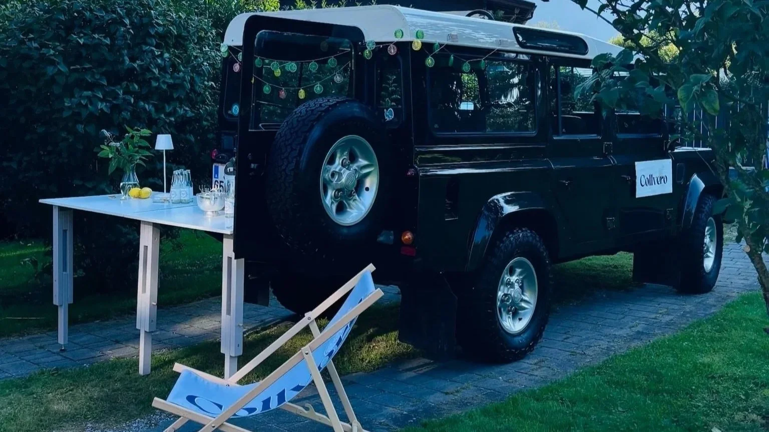 Ein schwarzer Geländewagen (Land Rover) steht auf einer gepflasterten Fläche in einem Garten. An der Seite des Wagens ist ein weißes Schild mit dem Schriftzug "Collvero". Vor dem Wagen steht ein weißer daumendicker Tisch mit Getränken und Geschirr, sowie eine flüssige Lichtquelle. Über dem Wagen hängen bunte Lichterketten. Im Vordergrund liegt ein abgestellter Liegestuhl mit blauer Auflage, auf dem steht "Sue".