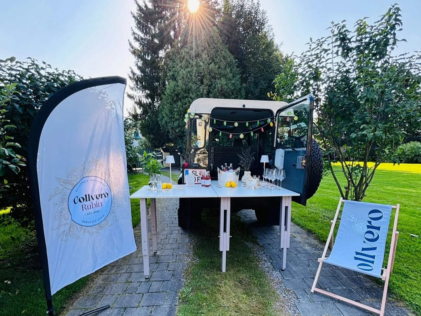 Ein mobiler Barwagen in einem Garten, dekoriert mit Lichterketten, mit einem weißen Tisch, auf dem Getränke und Gläser stehen. Es gibt eine Flagge mit dem Logo 'Collvero Rubin' und einen Lounge-Stuhl mit dem gleichen Logo.
