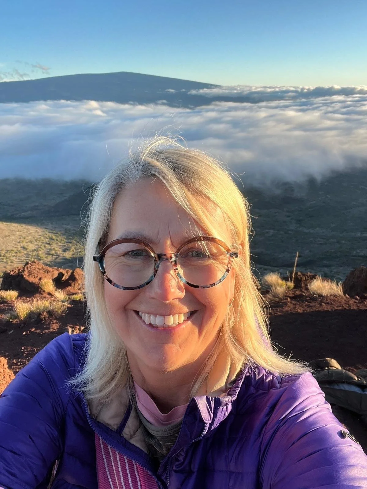 Frau mit Brille und blonder Haare lächelt in die Kamera vor einer Berglandschaft mit Wolken und blauem Himmel im Hintergrund.