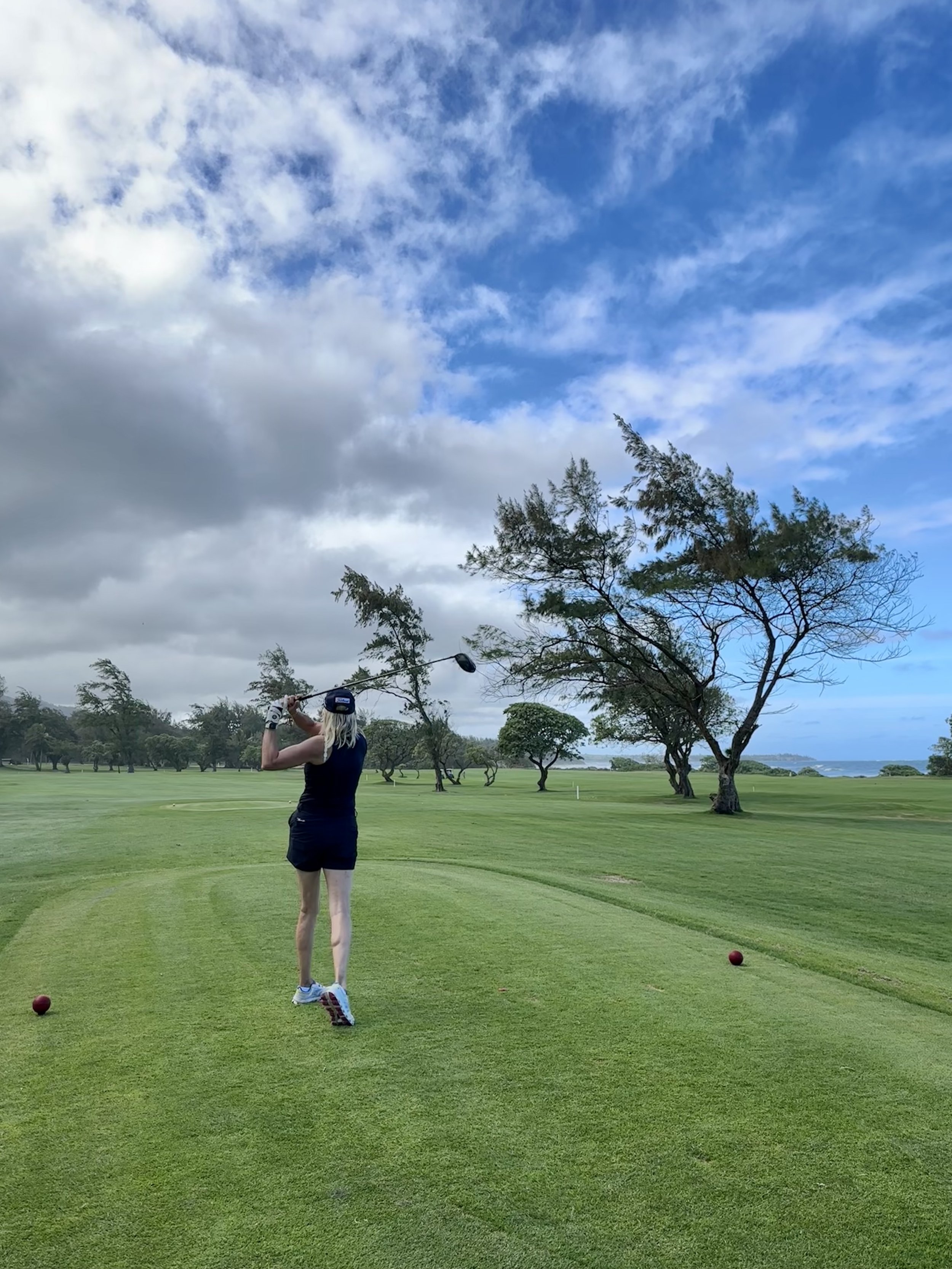 Person, die auf einem Golfplatz abschlägt, umgeben von Bäumen und bewölktem Himmel.