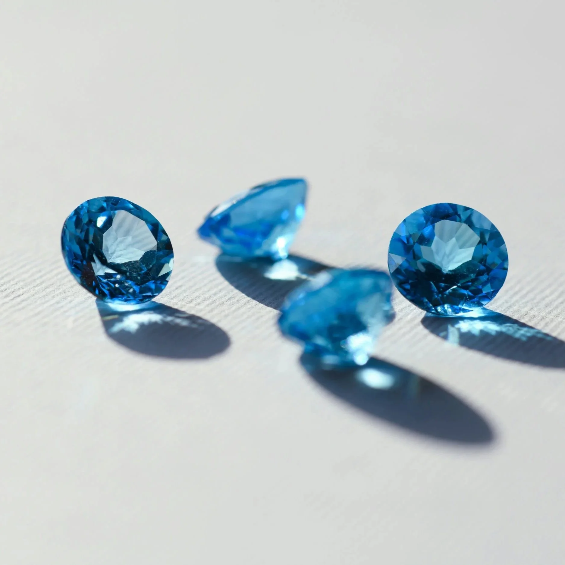 Trois pierres précieuses bleues facettées posées sur une surface blanche, avec des reflets et des ombres.