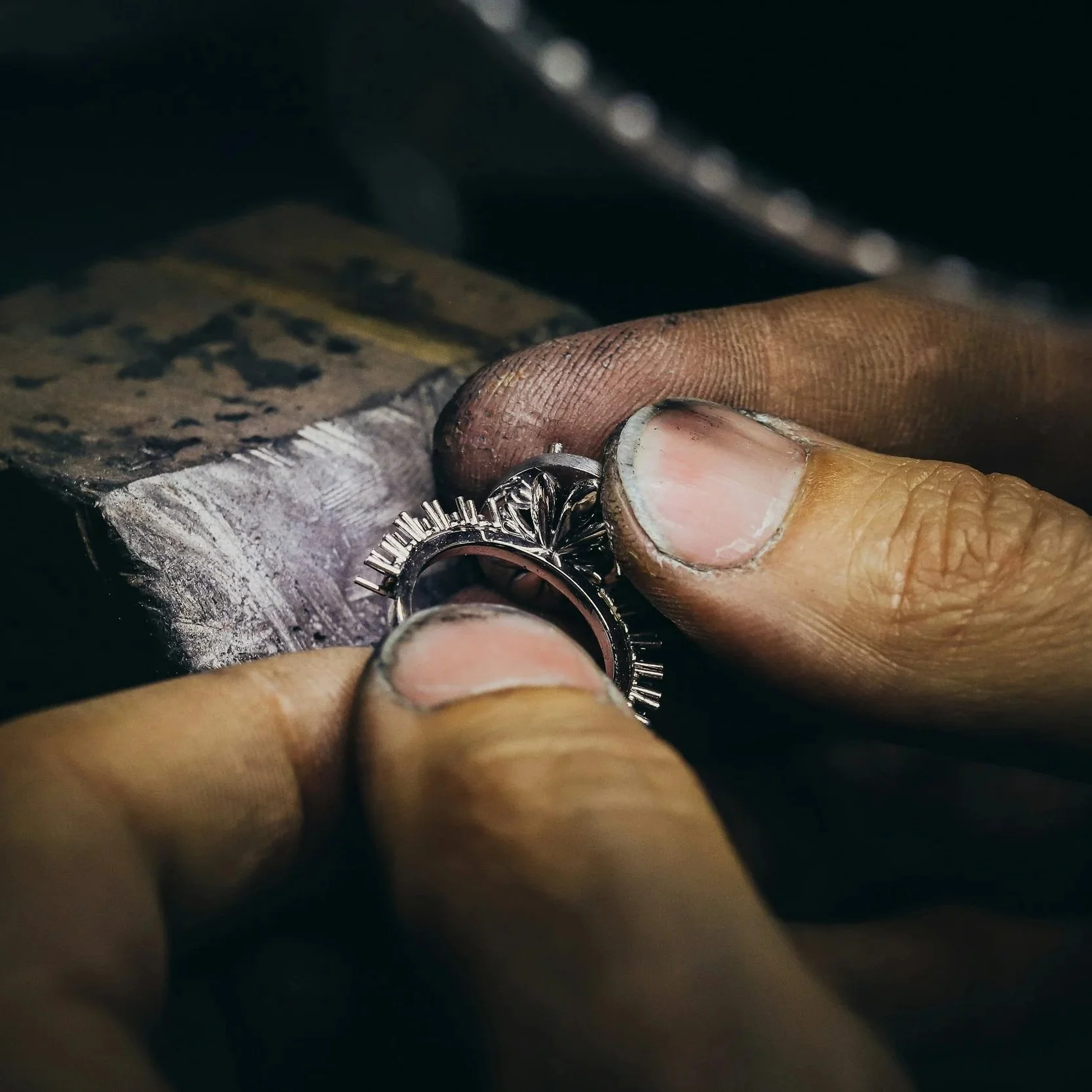 Une main tenant une bague en argent orné de détails complexes, dans un atelier de bijouterie, avec un banc de travail en arrière-plan.