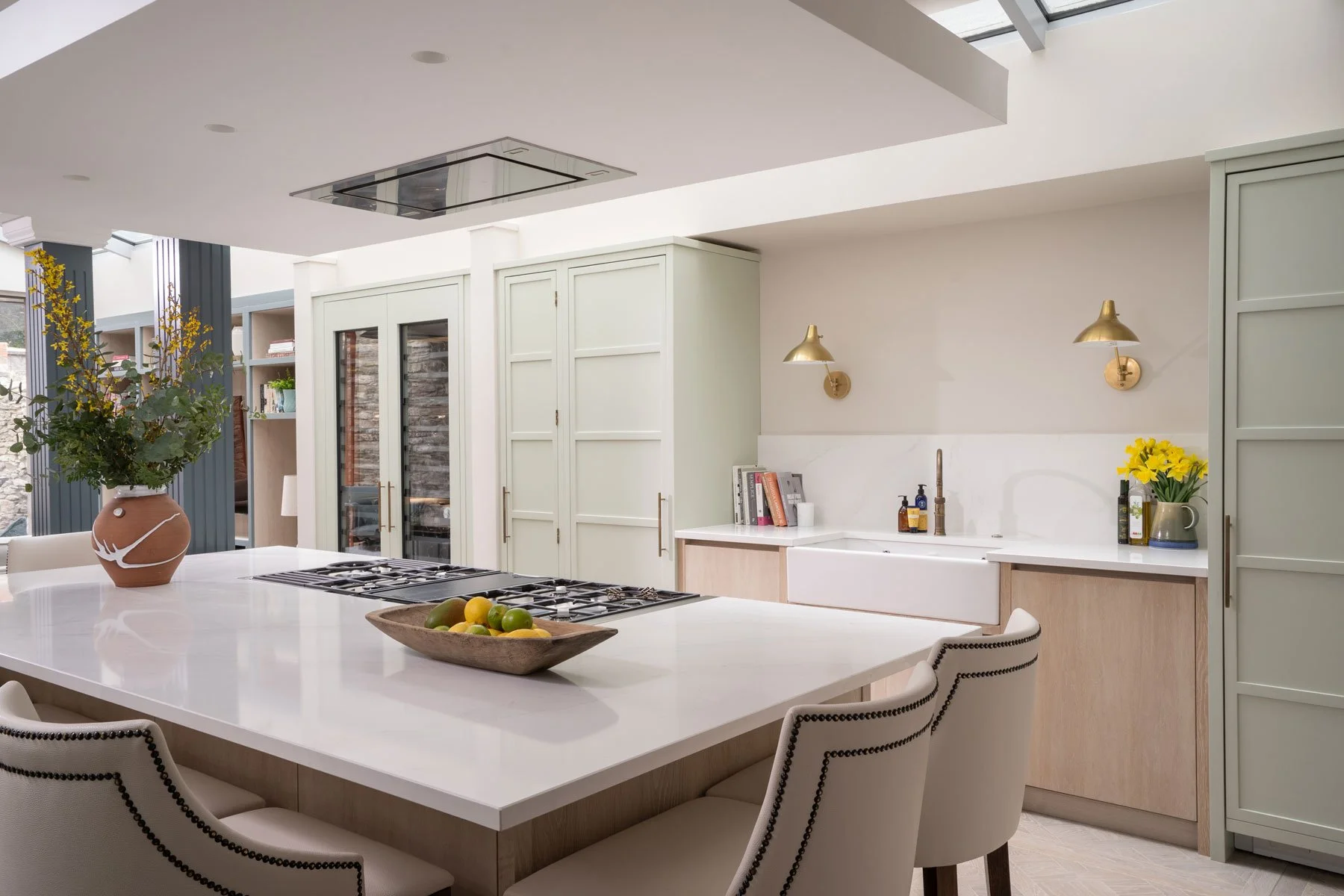 Asiko Architects Ltd — Modern kitchen with white island, vase with flowers, bowl of fruit, pendant lights, and built-in storage.