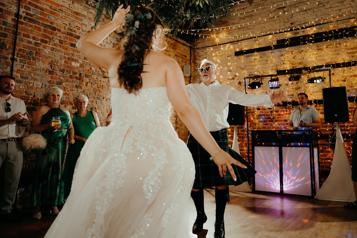 A bride in a white wedding dress and a person in a white shirt and kilt dancing at a wedding reception with guests watching in the background, brick wall decor, and string lights.