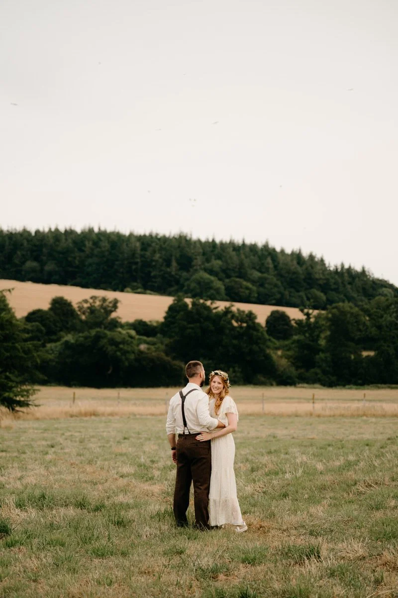 A couple standing in a grassy field, embracing and smiling at each other, with rolling hills and trees in the background during daytime.