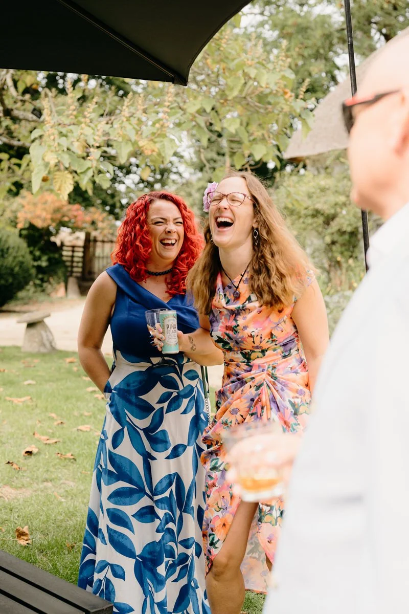 Two women laughing and enjoying drinks outdoors at a wedding in Dorset, with one woman wearing a blue and white dress and the other in a floral dress, under a black umbrella in a park-like setting.