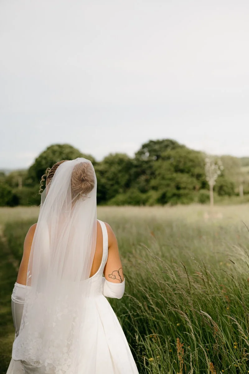 A woman dressed in a wedding gown and veil stands in a field of tall grass, facing away from the camera with trees in the background.