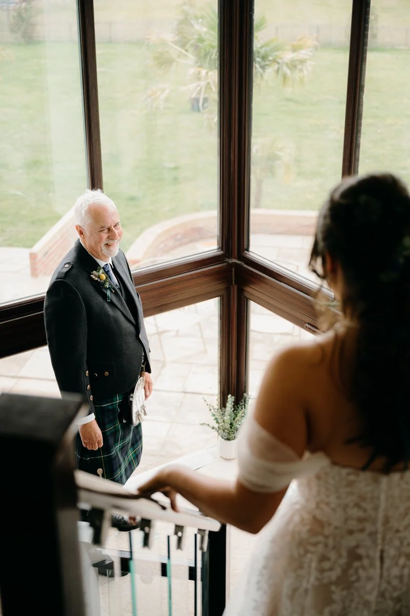 A bride looking down a staircase at an older man smiling in front of large windows with a greenery backyard view.