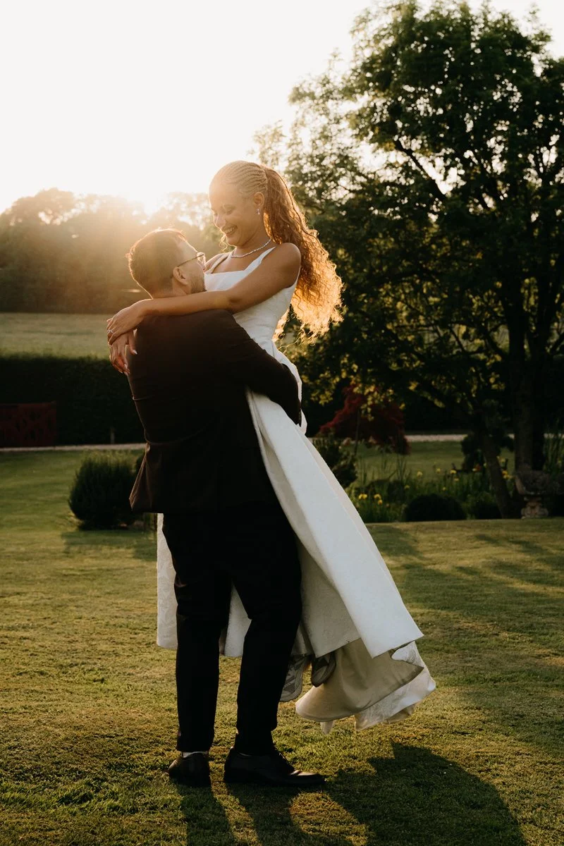 A man in a black suit lifting a woman in a white wedding dress outdoors at sunset, with trees in the background.