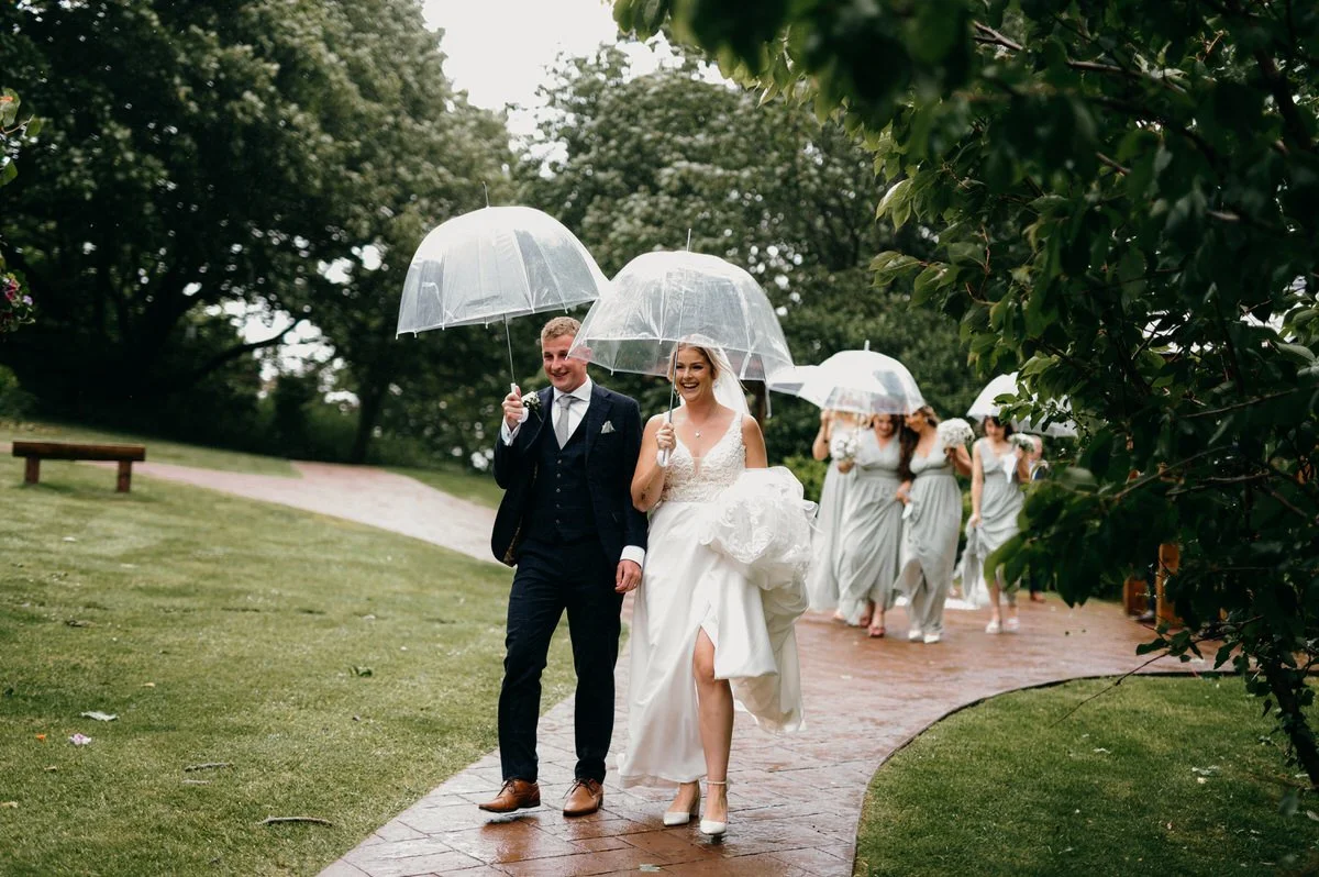 Bride and groom walking with bridesmaids under umbrellas on a rainy day in a garden.