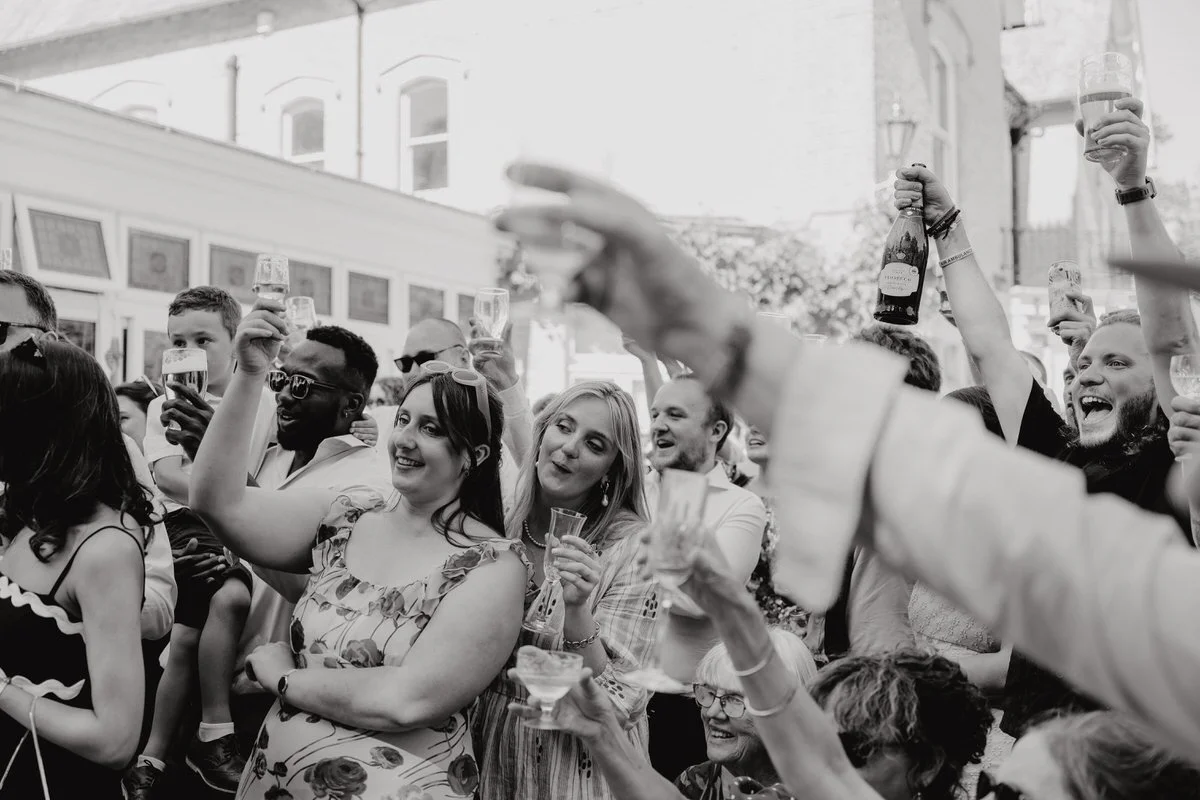 A crowd of people celebrating at an outdoor event, raising glasses of champagne and beer, smiling and enjoying each other's company. Photographed by Dorset wedding photographers pip&clo at an outdoor wedding ceremony at Radipole Manor Weymouth.