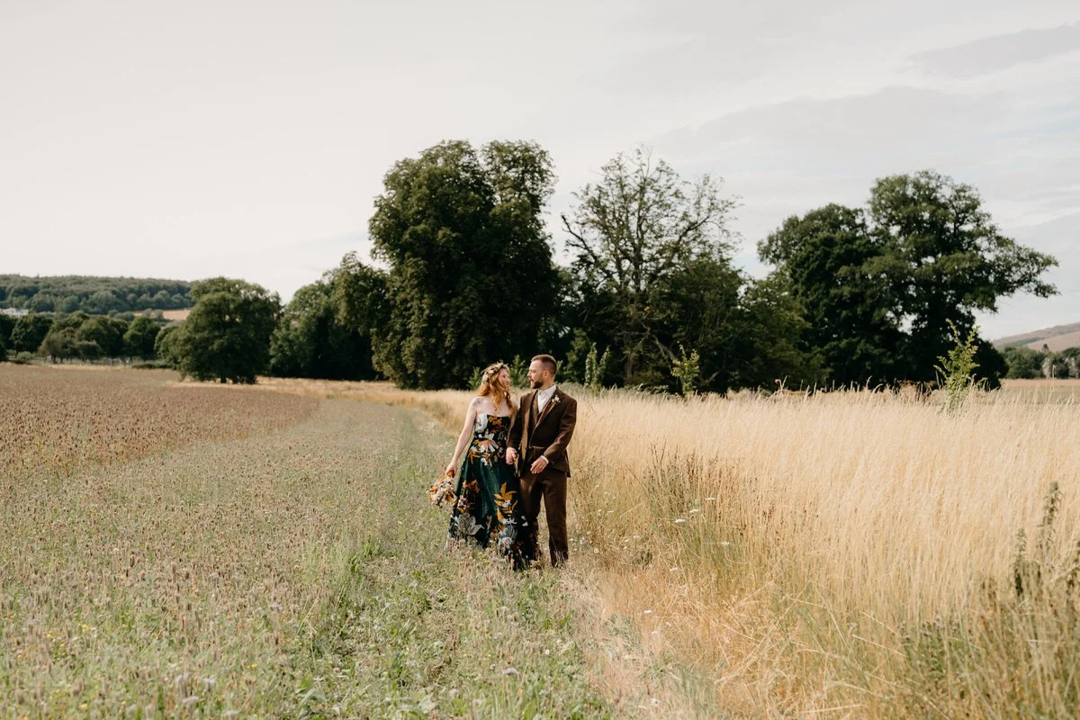 A married couple walking together in a field of tall grass on a cloudy day, holding hands and looking at each other photographed by Dorset wedding photographers pip&clo.
