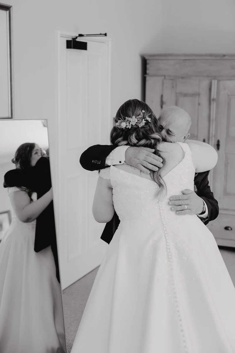 A bride in a wedding gown hugging an elderly man, likely her father, as her reflection is seen in a mirror.