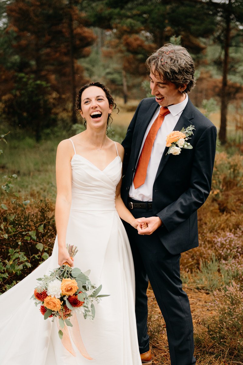 A happy bride and groom stand outdoors among trees, smiling and laughing. The bride wears a white wedding dress, holding a bouquet of orange and red flowers. The groom wears a dark suit with an orange tie and a boutonniere, holding the bride's hand.