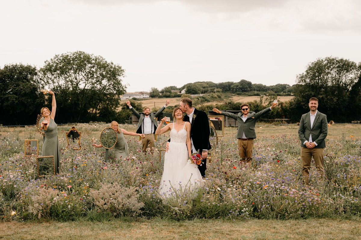 Wedding couple with bridal gown and tuxedo kissing in a flower field surrounded by friends in formal attire celebrating with drinks, some holding picture frames, on a cloudy day.