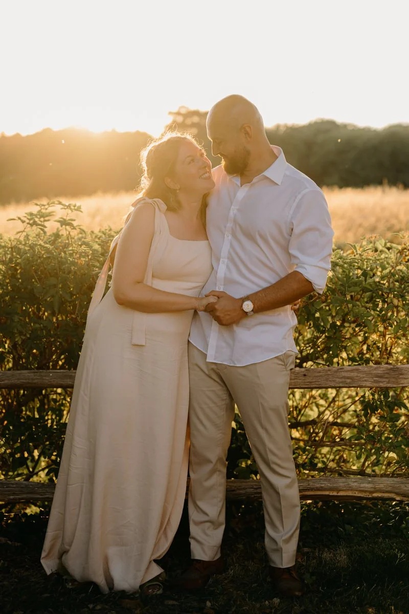 A wedding couple standing close together outdoors during sunset, smiling and holding hands, with a wooden fence and green foliage in the background. Golden Hour wedding photography in Dorset.