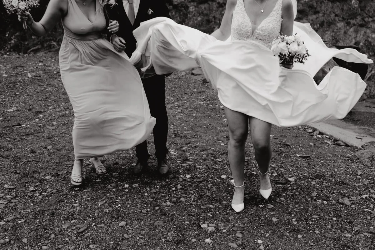 A black and white photo of a bride in a wedding dress holding a bouquet, a woman in a dress, and a man in a suit, with the bride being lifted by the other two.