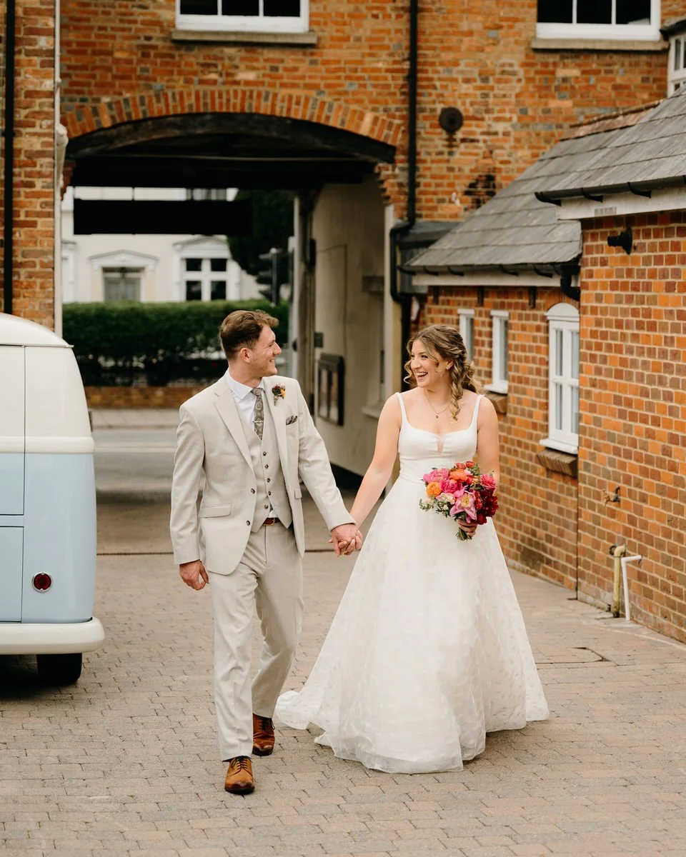 A bride and groom walking hand in hand outside a brick building, smiling at each other. The bride wears a white wedding dress and holds a colorful bouquet photographed by Dorset wedding photographers pip&clo at Wimborne Town Hall.