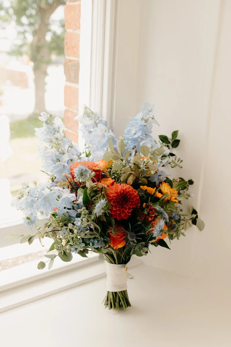 A bouquet of flowers with blue, orange, and white blooms sitting on a window sill indoors.