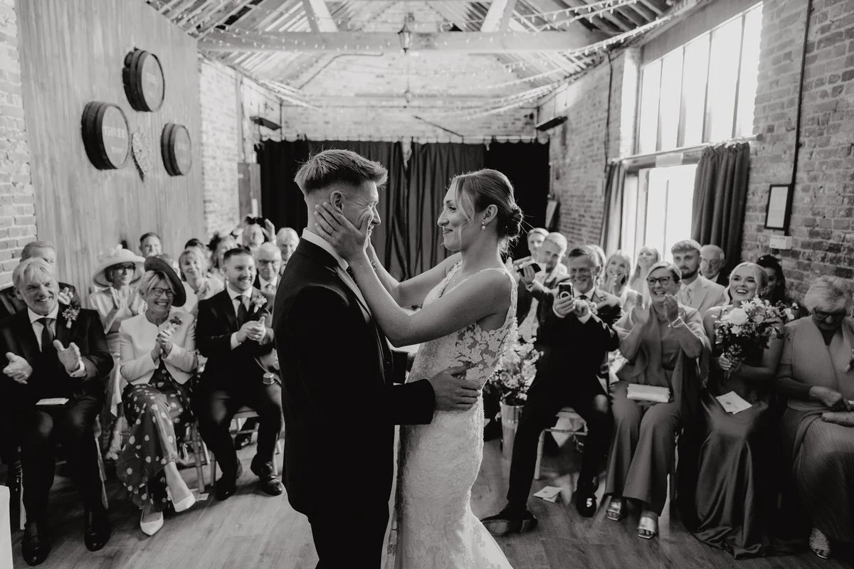 A bride and groom dance together during their wedding ceremony, surrounded by guests in a rustic indoor venue with brick walls and large windows.