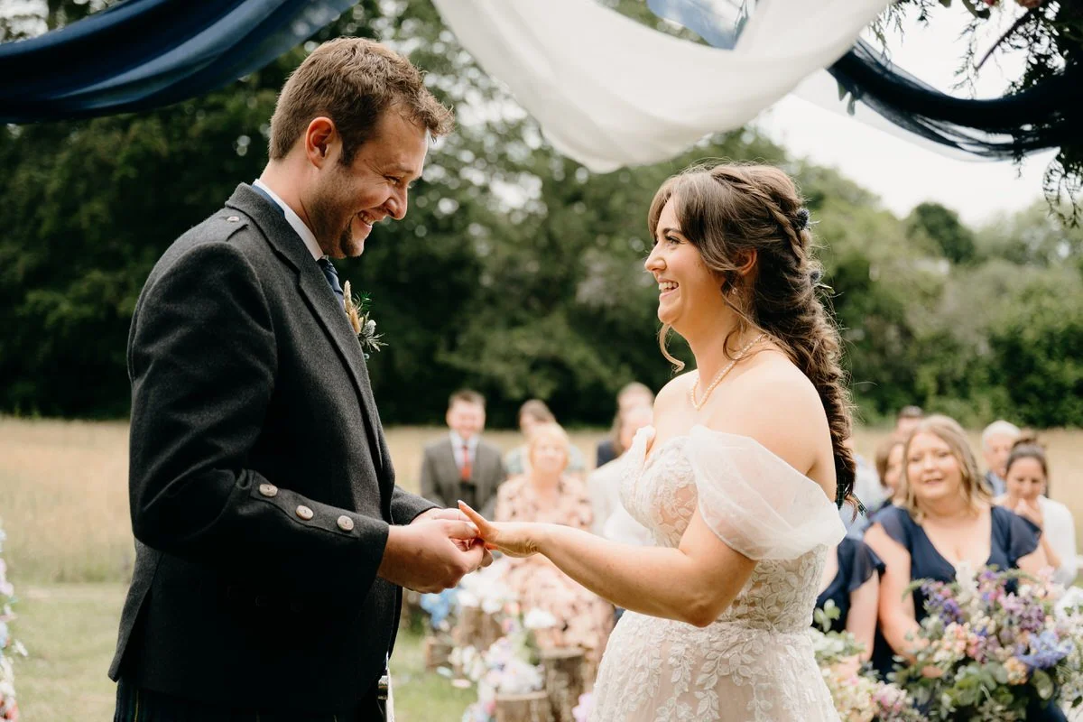 A bride and groom exchanging rings during an outdoor wedding ceremony, surrounded by guests and floral decorations.