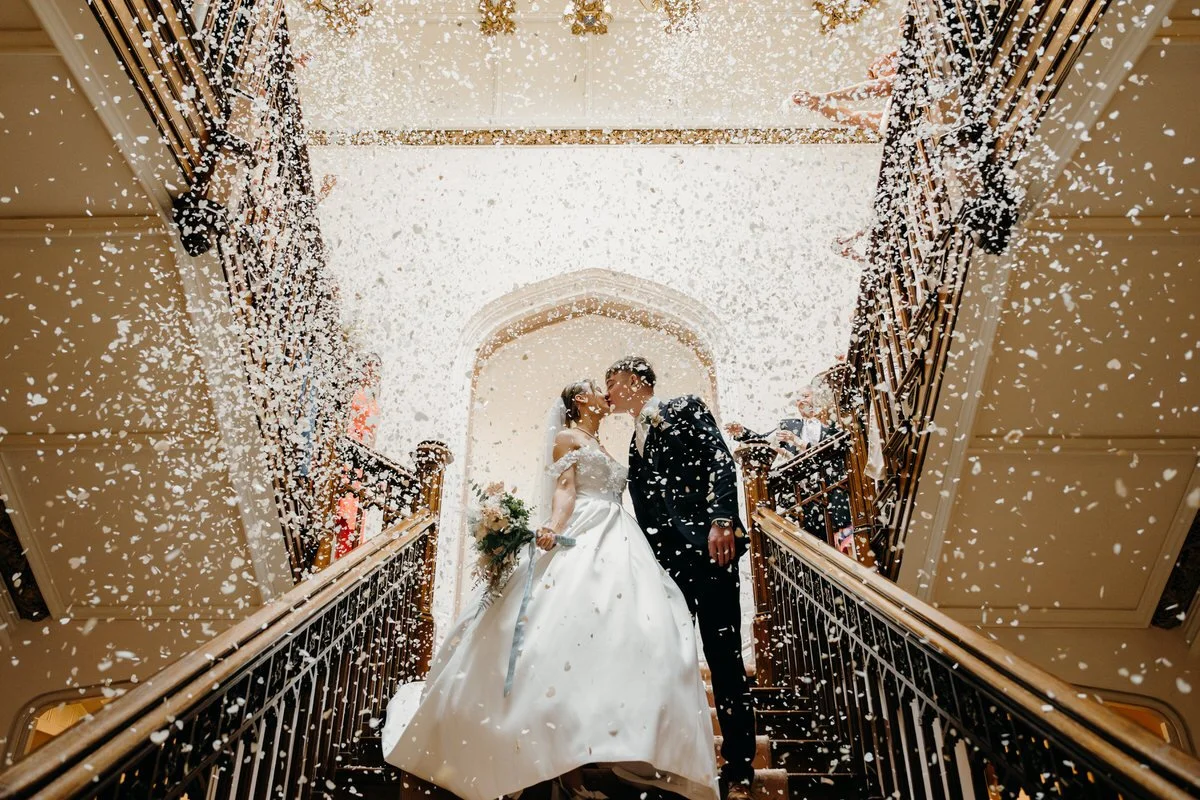 A bride and groom share a kiss on a staircase during their wedding, surrounded by falling confetti, with guests watching from the sides.