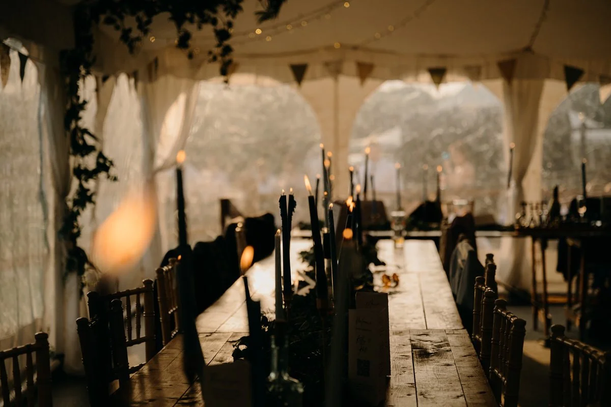 A decorated outdoor wedding marquee with a long wooden table and chairs, lit with candles and string lights, during sunset.