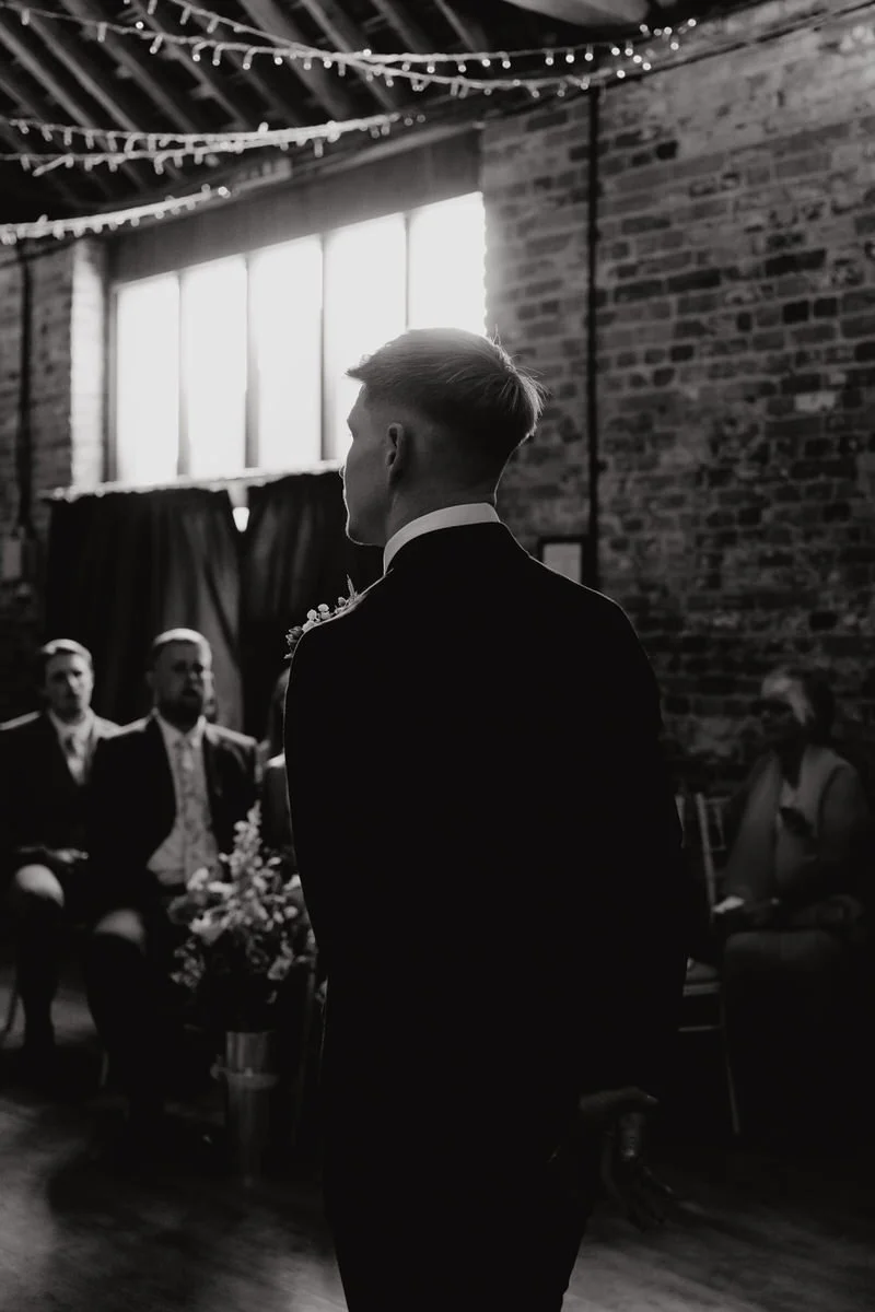 Black and white photo of a groom standing during a wedding ceremony in an industrial-style venue with exposed brick walls, large windows, and string lights overhead, with guests seated and watching.