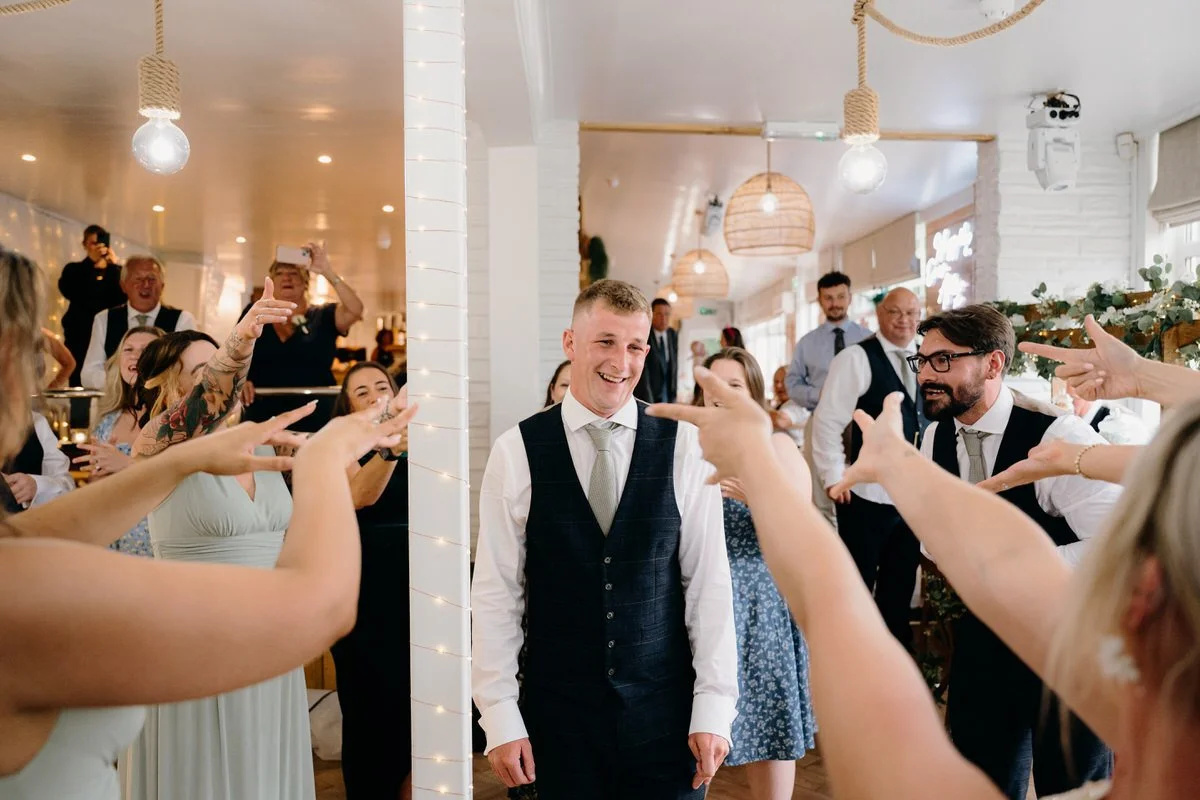 Groom in a suit smiling as friends and family celebrate and point at him during wedding reception.