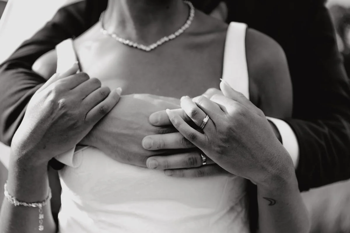 Close-up of a married couple touching their hands, with the woman wearing a pearl necklace and wedding rings, in black and white.