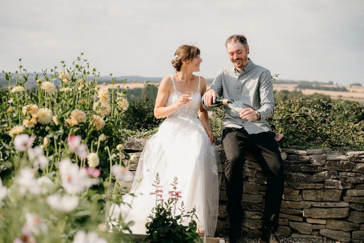A couple in wedding attire celebrating outdoors with champagne, sitting on a stone wall surrounded by flowers and greenery, with a landscape view in the background.